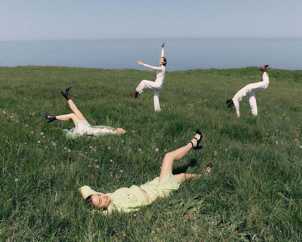 Four women in a grassy field near the ocean. Two are lying down, and two are striking poses, all wearing light-colored clothing.