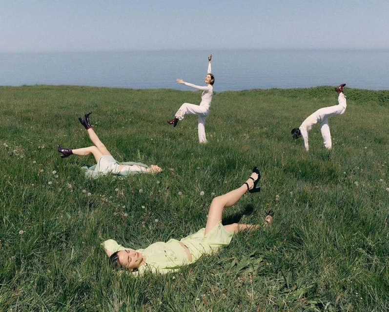 Four women in a grassy field near the ocean. Two are lying down, and two are striking poses, all wearing light-colored clothing.
