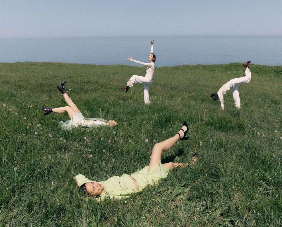 Four women in a grassy field near the ocean. Two are lying down, and two are striking poses, all wearing light-colored clothing.