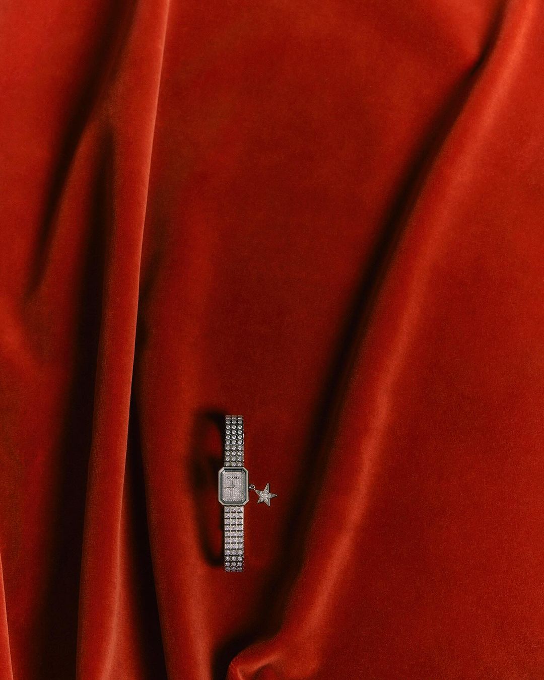 Close-up of a silver wristwatch with charm, against a backdrop of draped, textured orange velvet fabric.