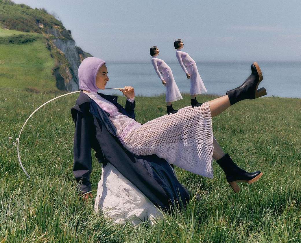 A woman in a lilac hooded dress and black boots sits on a rock in a green field by the sea, with two clone figures behind her.