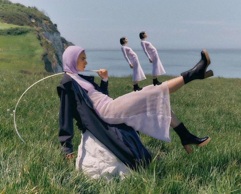 A woman in a lilac hooded dress and black boots sits on a rock in a green field by the sea, with two clone figures behind her.