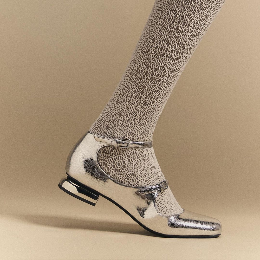 Close-up shot of a leg wearing beige lace stocking and a shiny silver Mary Jane heel with an ankle strap, set against a beige background.
