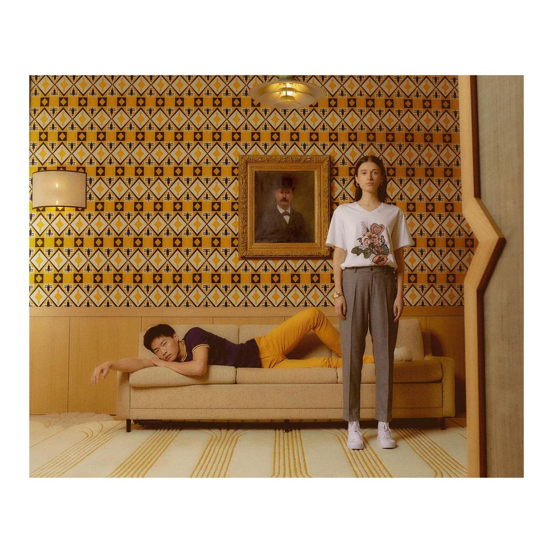 Indoor fashion shot featuring an Asian man reclining on a sofa and a Caucasian woman standing beside him in a room with patterned wallpaper.