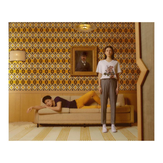 Indoor fashion shot featuring an Asian man reclining on a sofa and a Caucasian woman standing beside him in a room with patterned wallpaper.