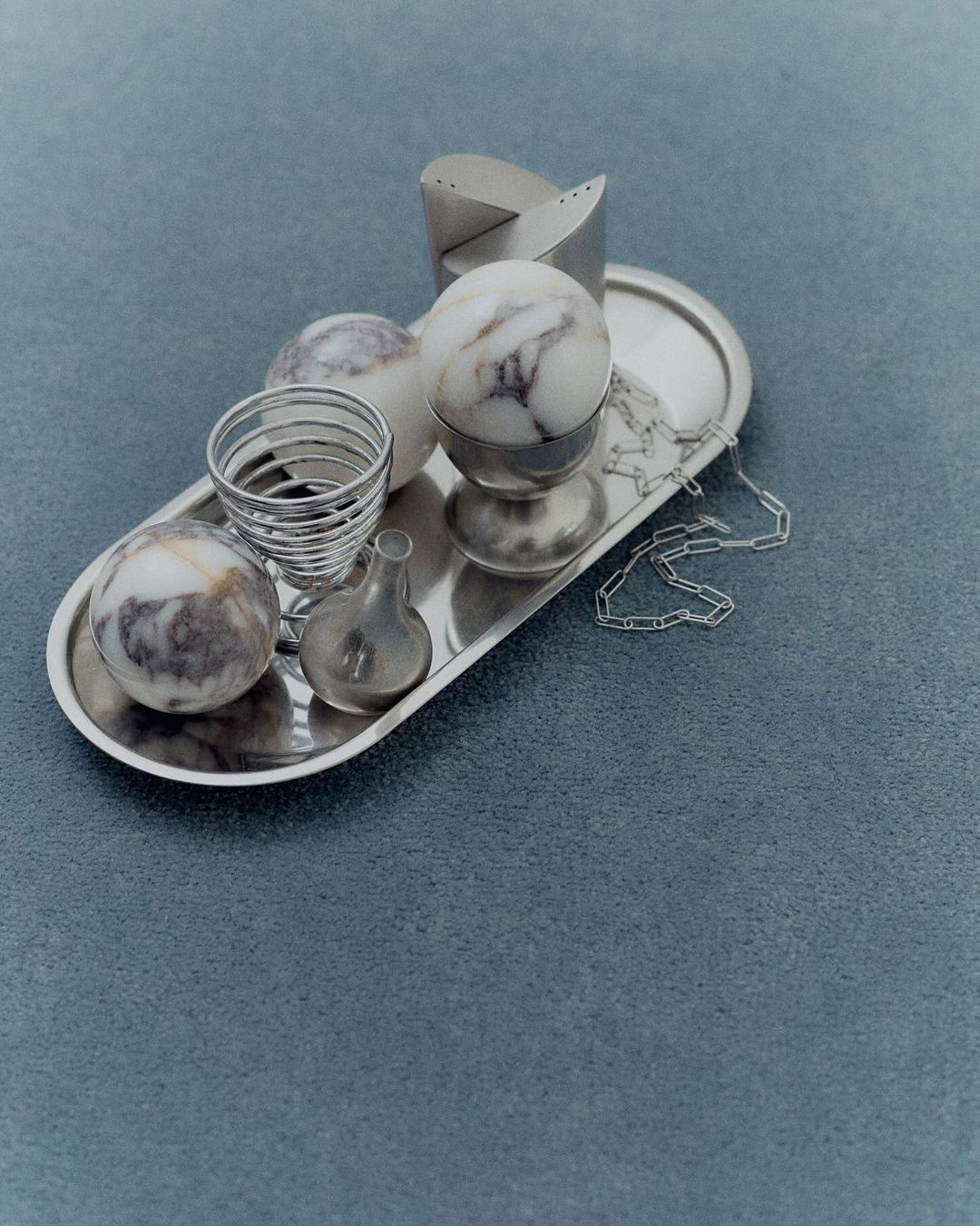 Still life with silver tray holding decorative spheres, a metal egg cup, and a chain, set against a light blue textured background.