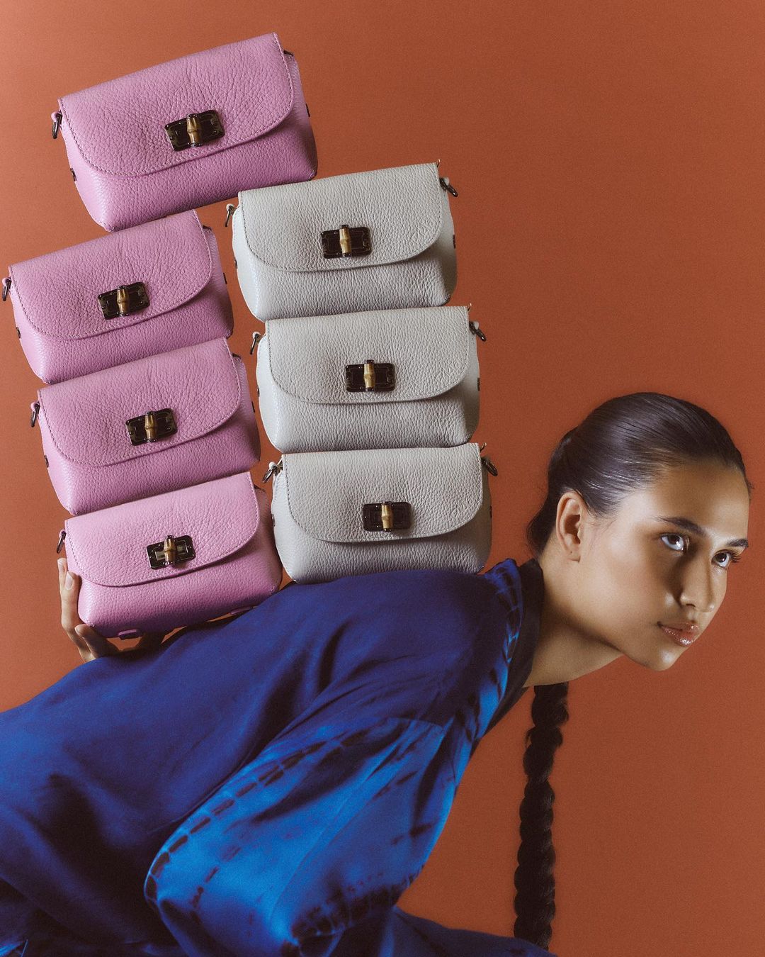 Fashion shot of a young woman with a stack of pink and gray leather purses balanced on her shoulder in a studio setting with an orange background.