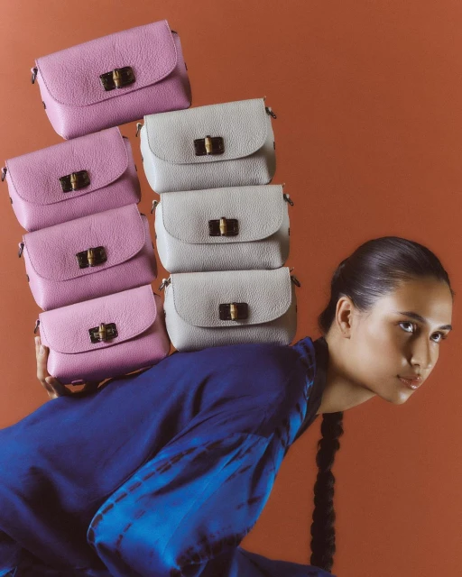 Fashion shot of a young woman with a stack of pink and gray leather purses balanced on her shoulder in a studio setting with an orange background.
