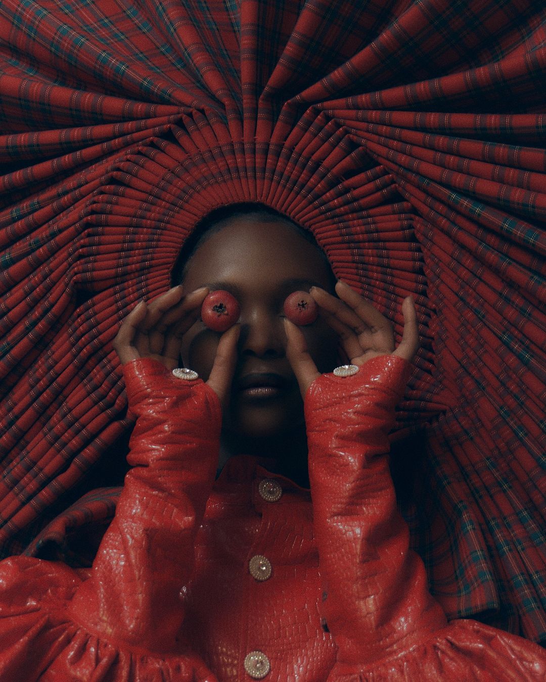 Woman with dark skin poses wearing a red plaid hat and dress, holding red fruit to her eyes in a studio shot. 