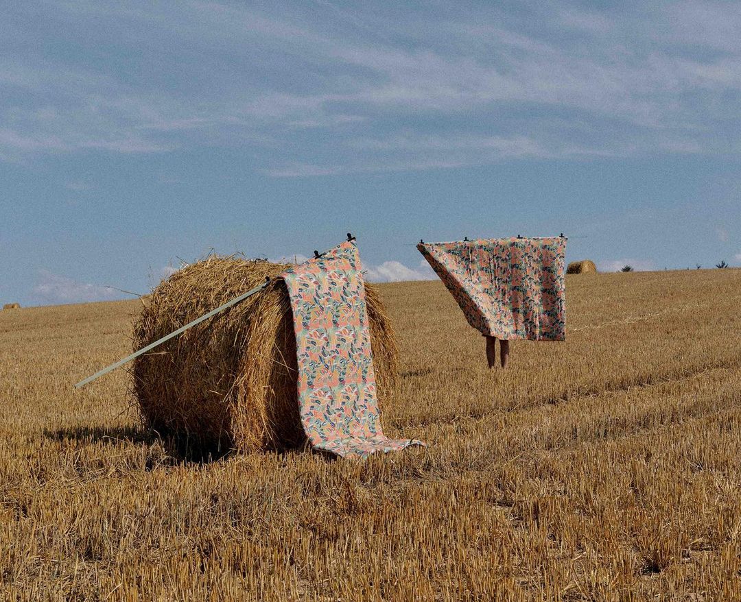 Two fabrics hang on a line between hay bales in a harvested wheat field under a bright blue sky during daytime, rural setting.