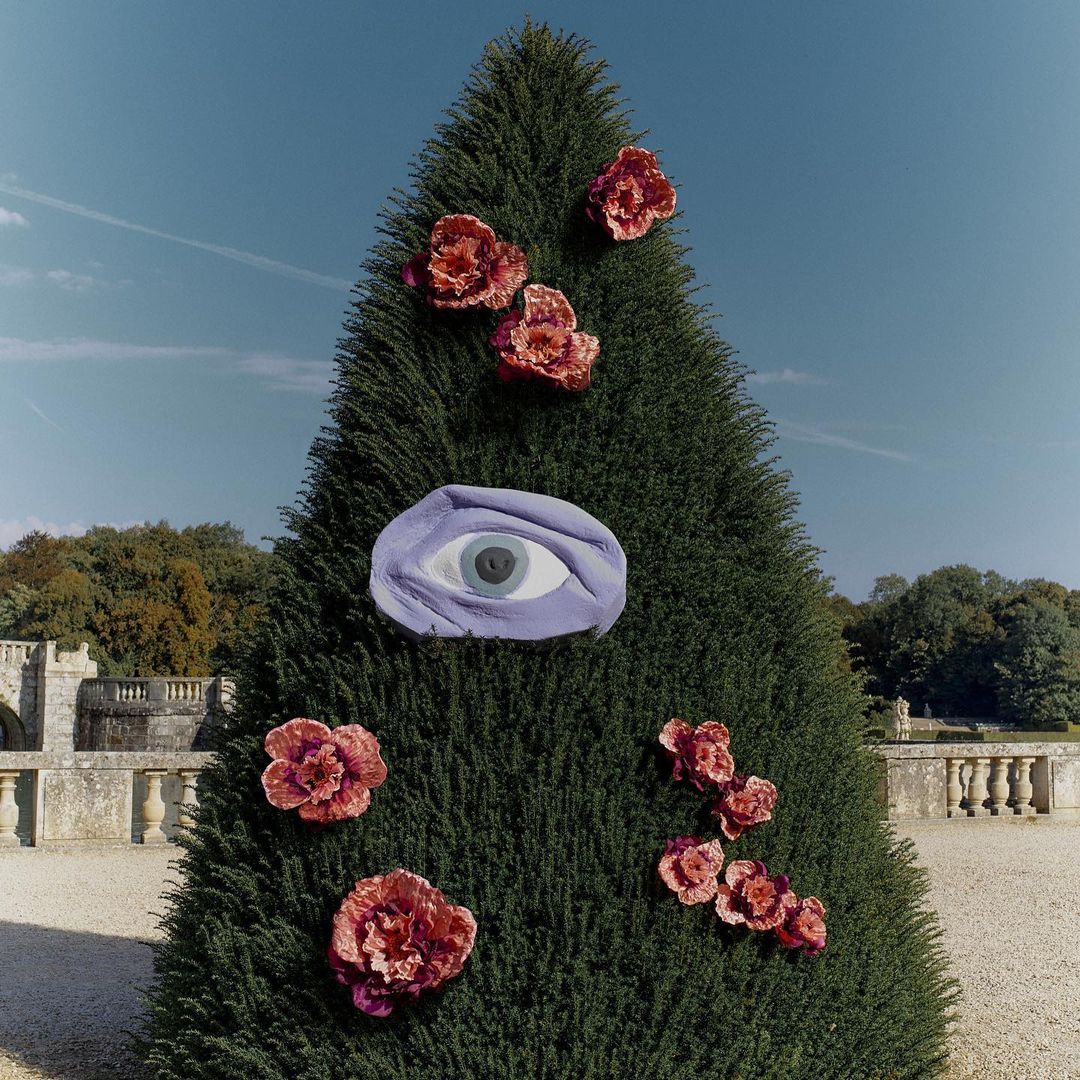 Conical topiary in an outdoor garden setting, adorned with pink fabric flowers and a large blue eye, set against a blue sky backdrop.