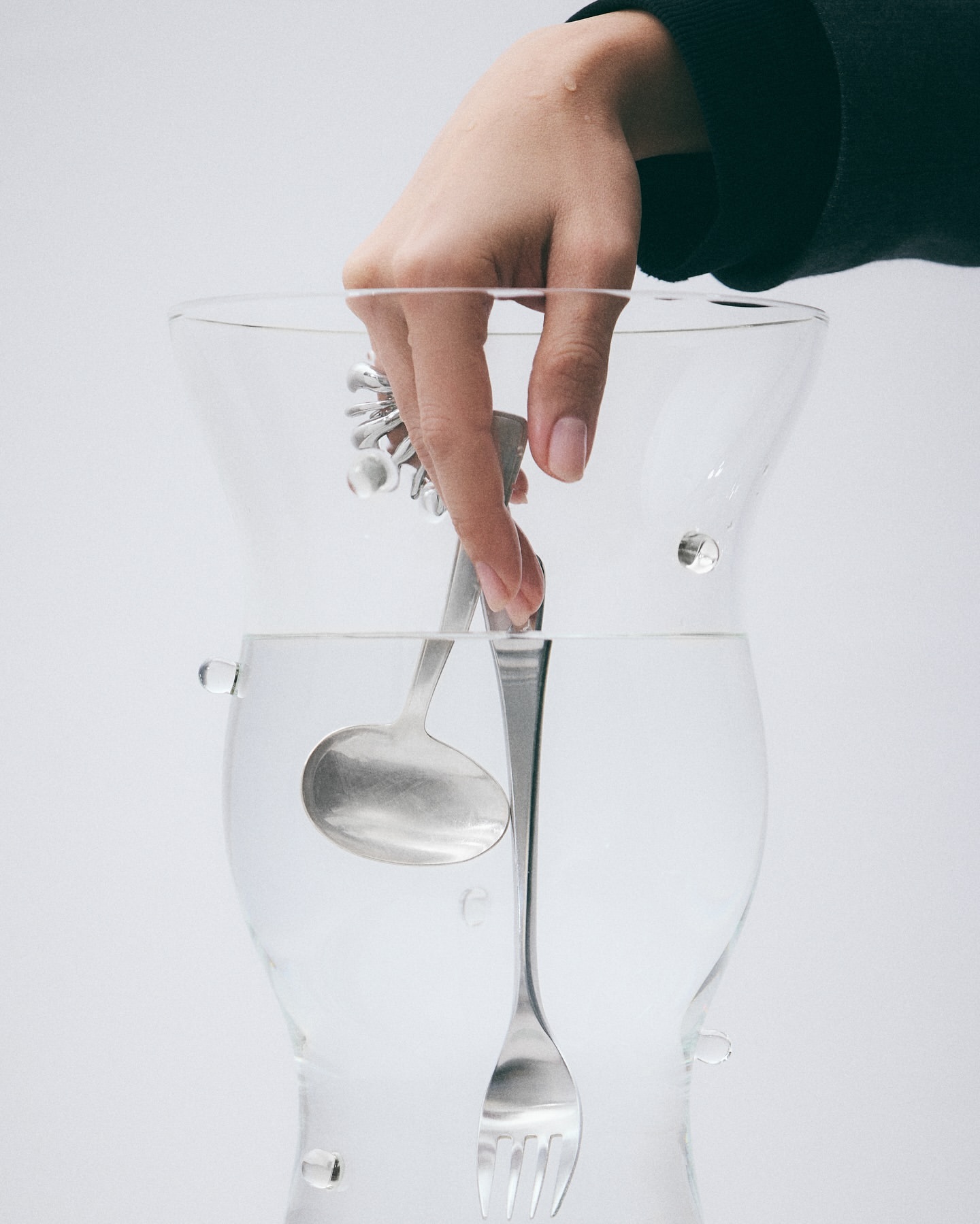 Hand places metal serving utensils – ladle, fork, tongs – inside clear glass container filled with water against white background.