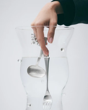 Hand places metal serving utensils – ladle, fork, tongs – inside clear glass container filled with water against white background.