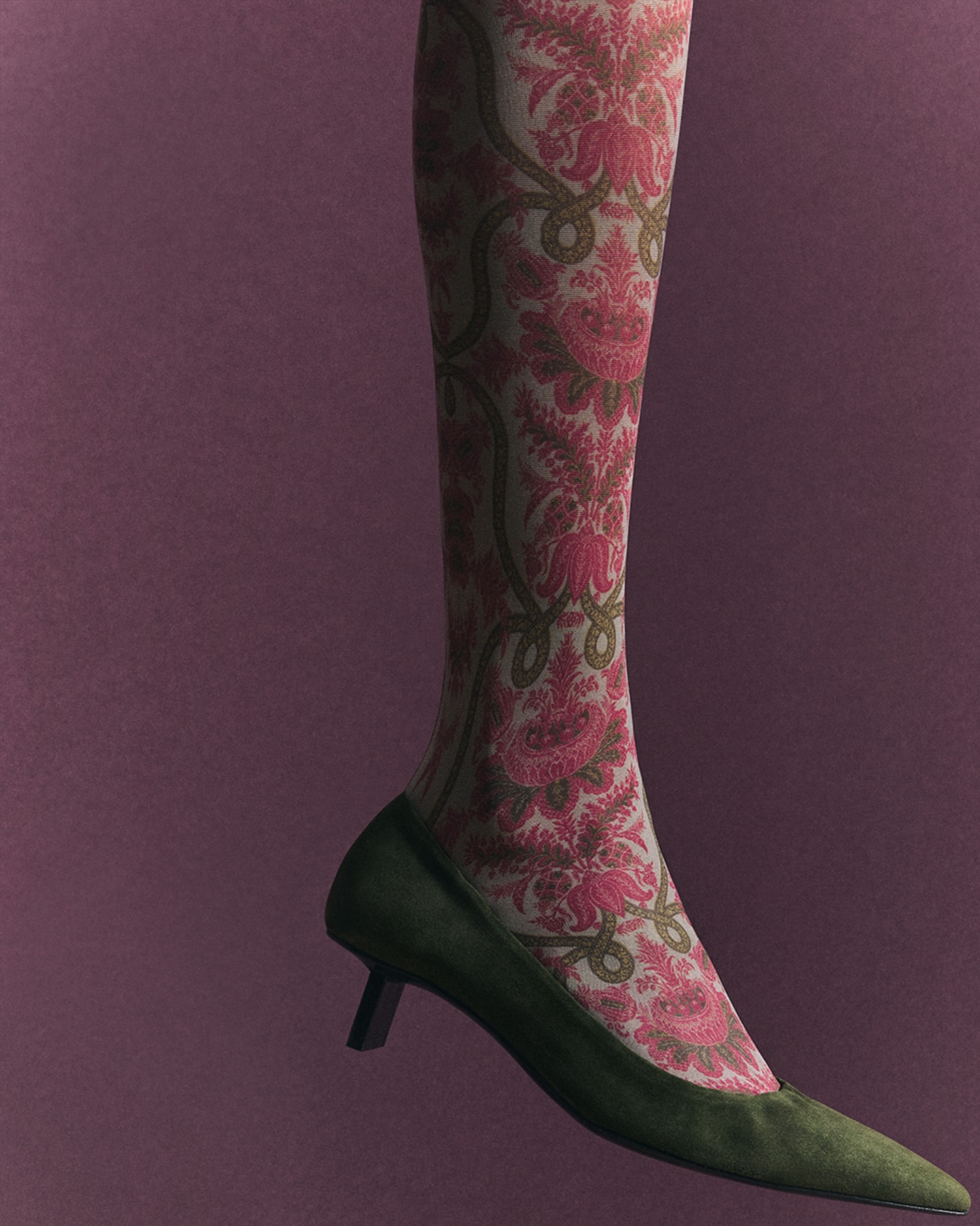 Close-up of a person's leg wearing patterned hosiery and a green kitten heel shoe against a mauve backdrop in a studio setting.