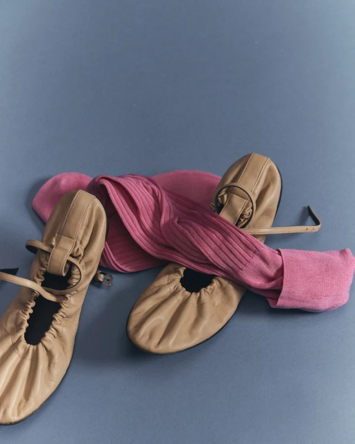 Still life studio shot of a pair of beige leather Mary Jane shoes alongside a pink ribbed knit scarf on a neutral grey-blue backdrop.