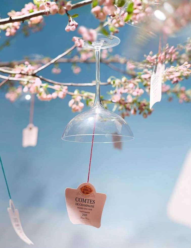 Upside-down wine glass suspended from a cherry blossom tree branch with tag, against a blue sky, creating a whimsical and artistic scene.