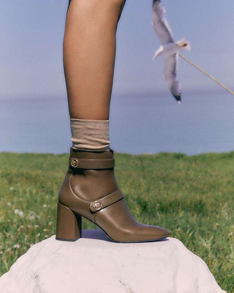 Fashion forward brown leather ankle boot with straps and block heel, displayed outdoors with grassy background and white stone.