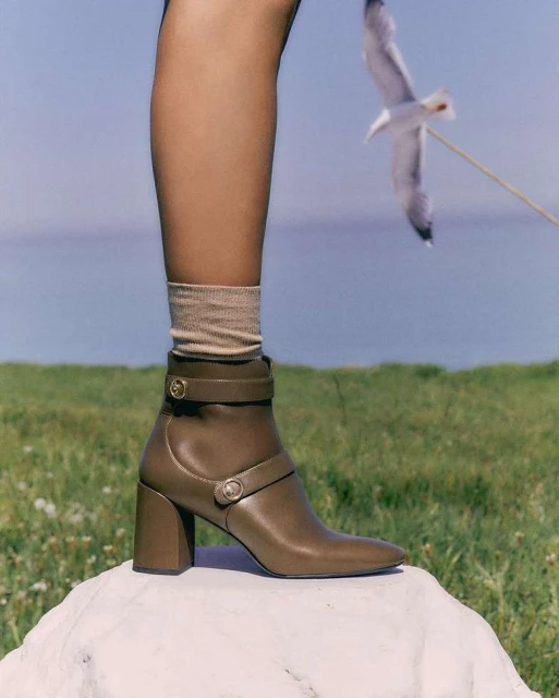 Fashion forward brown leather ankle boot with straps and block heel, displayed outdoors with grassy background and white stone.