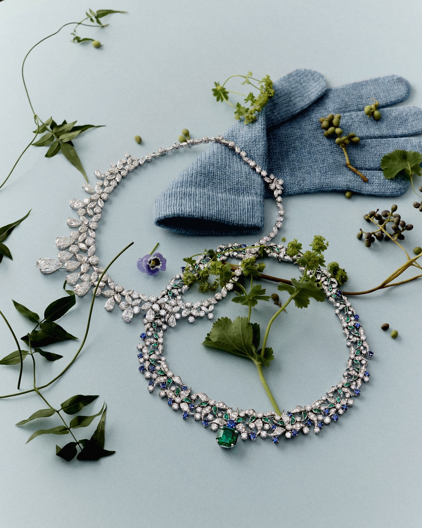 Still life of two gemstone necklaces, a knitted glove, and greenery on a light blue background in an indoor fashion product shot.