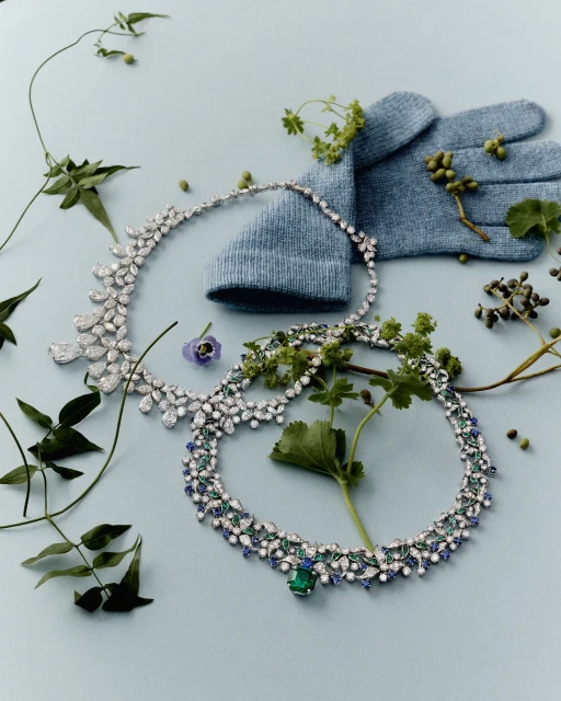 Still life of two gemstone necklaces, a knitted glove, and greenery on a light blue background in an indoor fashion product shot.