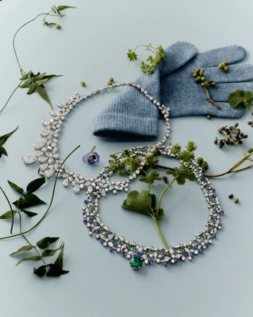 Still life of two gemstone necklaces, a knitted glove, and greenery on a light blue background in an indoor fashion product shot.