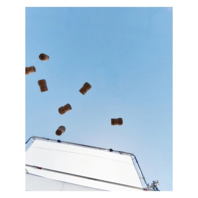 Several champagne corks are suspended mid-air against a bright blue sky, with a white scrim or reflector positioned below.
