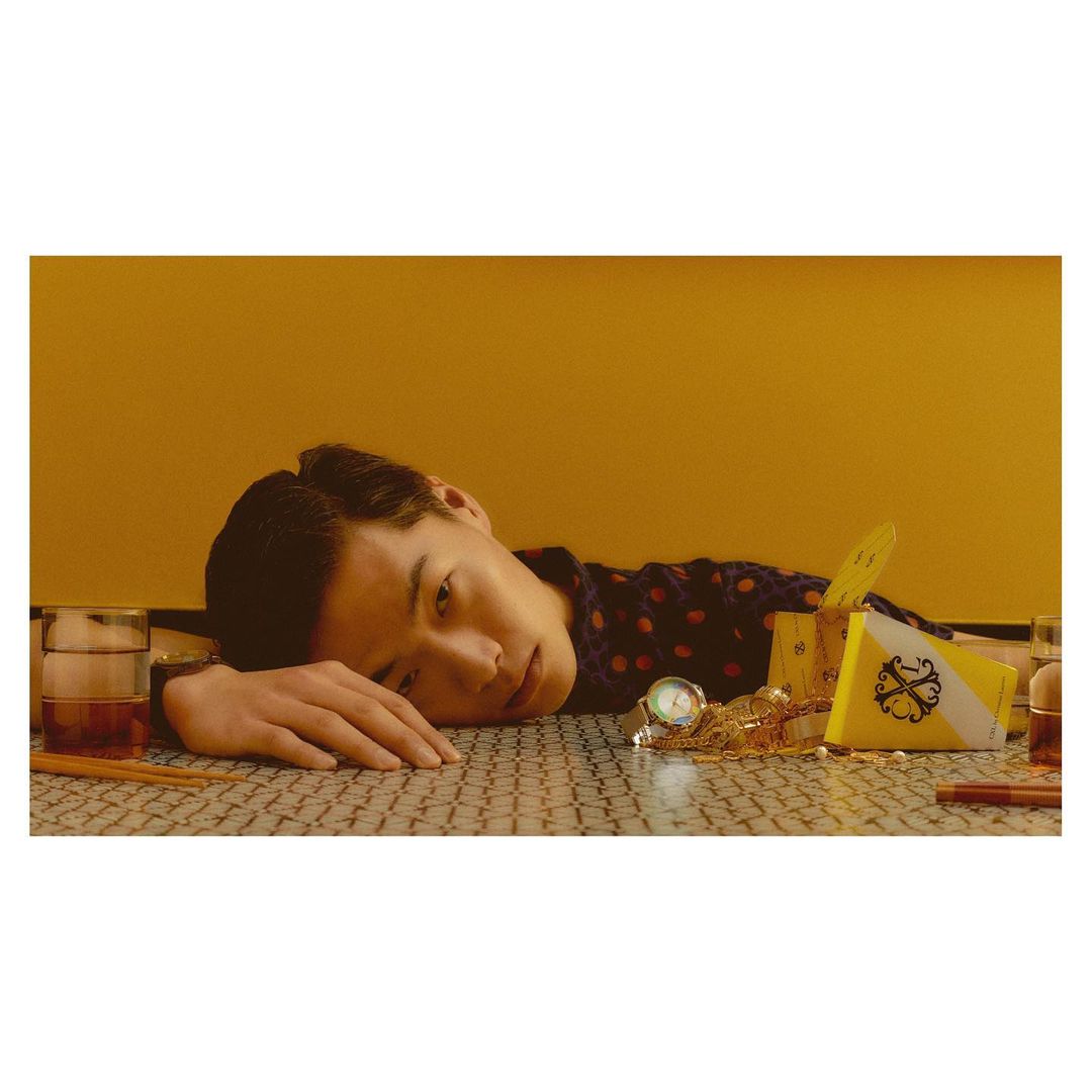 Asian man rests on a patterned table with gold jewelry, a wristwatch, and a glass of dark liquor with a yellow background.
