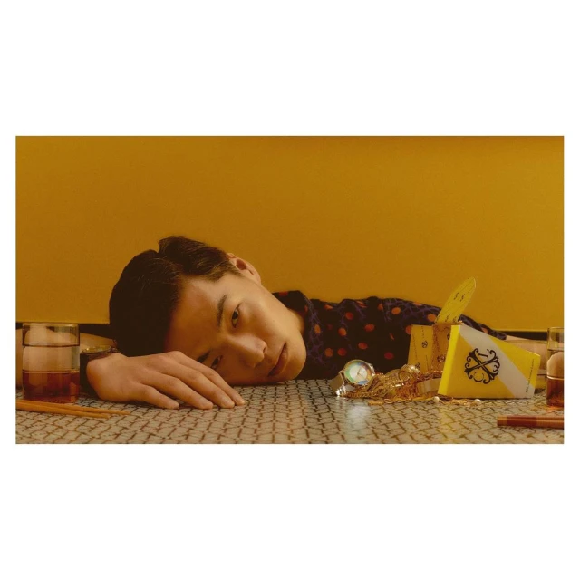 Asian man rests on a patterned table with gold jewelry, a wristwatch, and a glass of dark liquor with a yellow background.