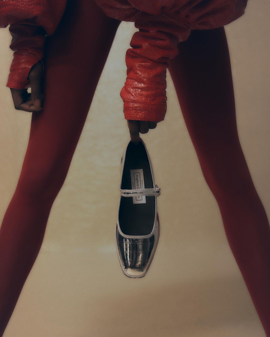 Fashion shot featuring an unrecognizable model wearing a red jacket and tights, holding a silver high-heeled shoe against a light backdrop.