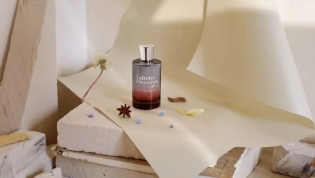 A perfume bottle surrounded by small botanicals on stacked stone blocks and curved paper sheets.