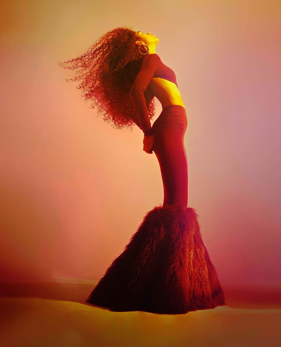 Adult woman with curly hair in cropped top and flared, fur-cuffed trousers bends backward in studio with gradient orange to lavender background.