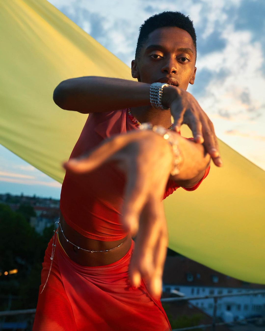 A young Black man poses, wearing a red mesh top and skirt with jewelry, against a yellow backdrop with sky in an outdoor setting.