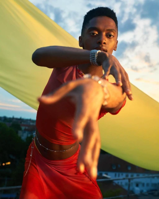 A young Black man poses, wearing a red mesh top and skirt with jewelry, against a yellow backdrop with sky in an outdoor setting.