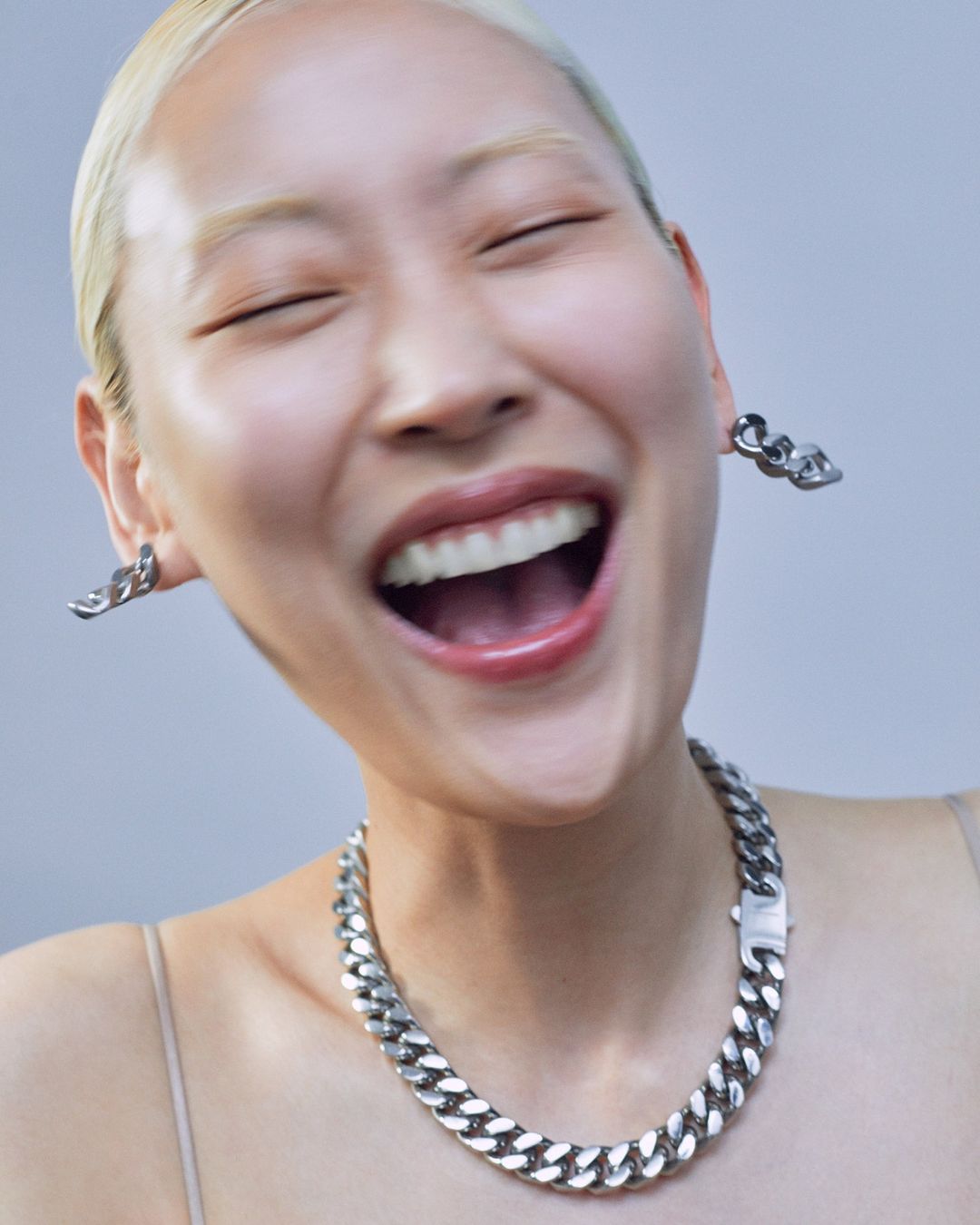 Close-up of a smiling Asian woman with short blonde hair, wearing chain jewelry, against a soft gray background.