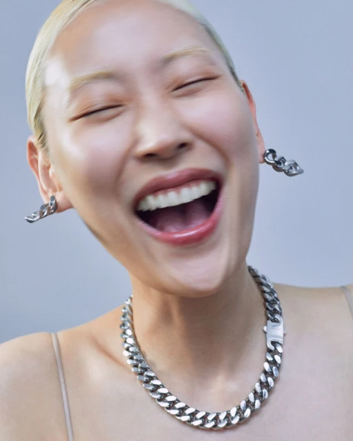 Close-up of a smiling Asian woman with short blonde hair, wearing chain jewelry, against a soft gray background.