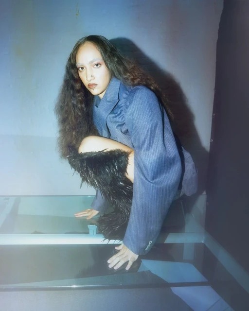 Woman in blue pinstripe blazer poses in a squatted position with dark feather-trimmed leg coverings on a glossy surface, studio lighting.
