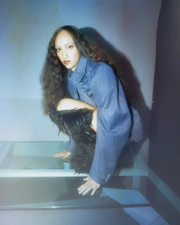 Woman in blue pinstripe blazer poses in a squatted position with dark feather-trimmed leg coverings on a glossy surface, studio lighting.