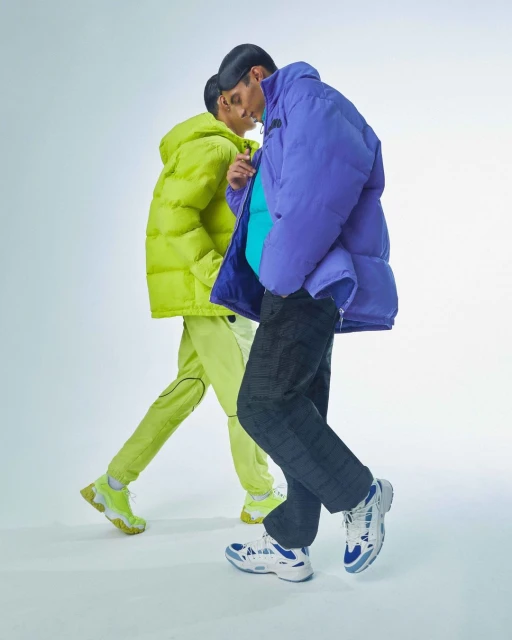 Two people in puffy coats walking against a light backdrop. One person wears a purple jacket and the other wears a bright lime-green jacket.