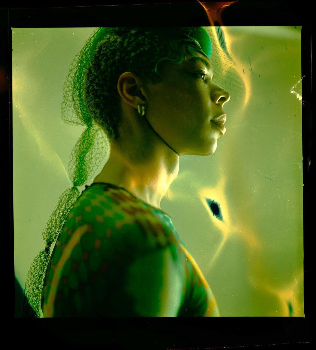Portrait of a woman in profile wearing a patterned top and netting in a studio with green and yellow lighting, shot with shallow depth of field.