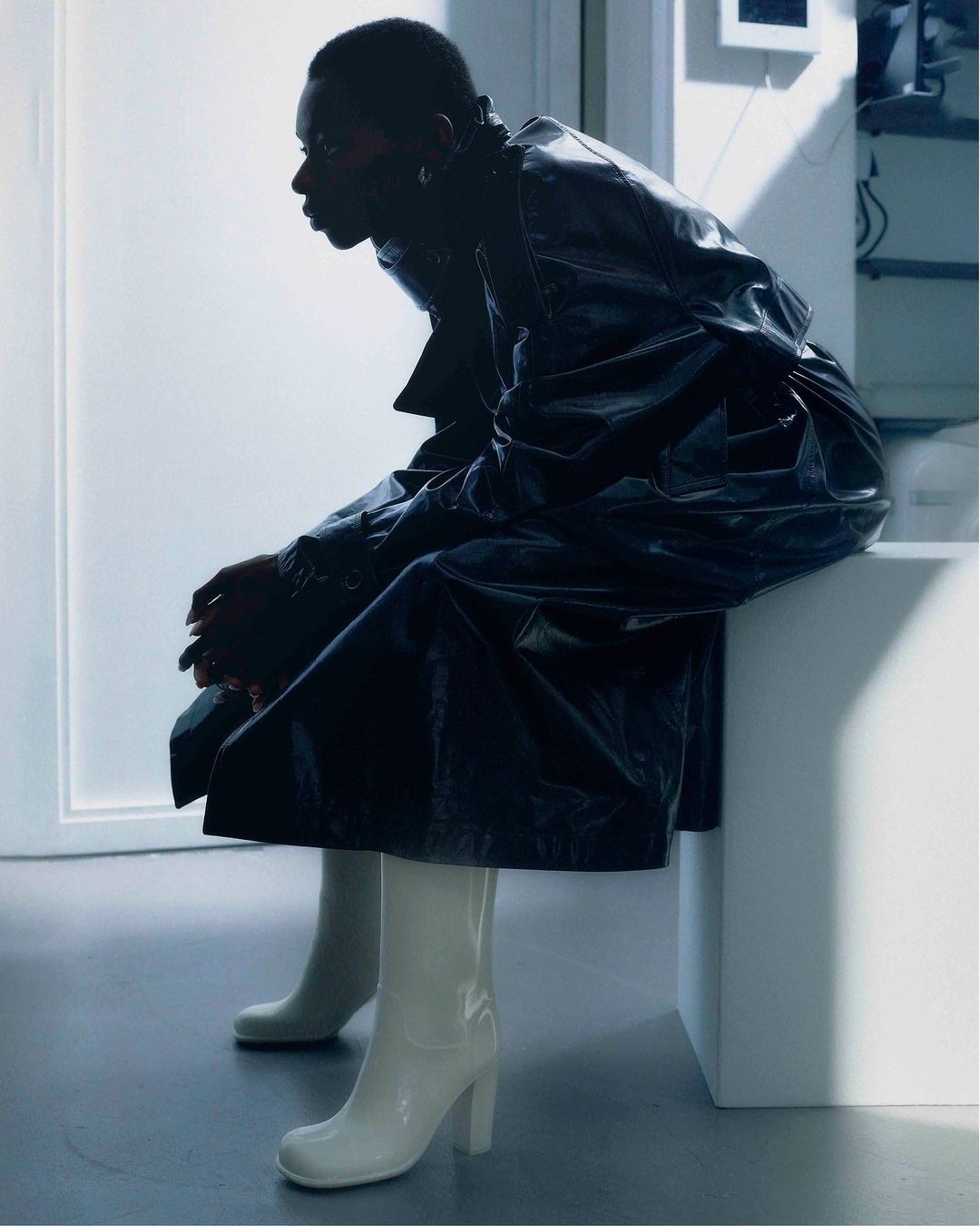 An African-American woman in glossy coat and white boots sits indoors, side profile, with soft light highlighting fashion and minimalist aesthetic.