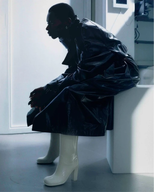 An African-American woman in glossy coat and white boots sits indoors, side profile, with soft light highlighting fashion and minimalist aesthetic.