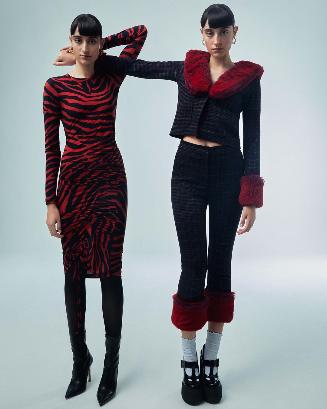 Two women pose in a photo studio; one wears a red tiger-striped dress, and the other wears a grey jacket and pants with red faux fur trim.