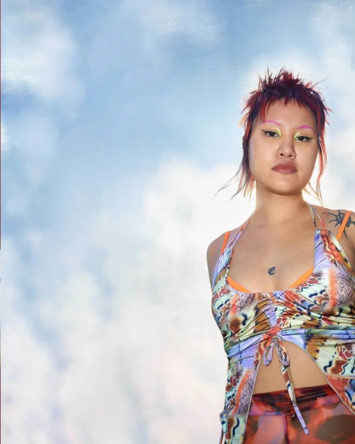 Low-angle portrait of a stylish Asian woman with colorful makeup and butterfly top against a cloudy blue sky, looking at the camera.