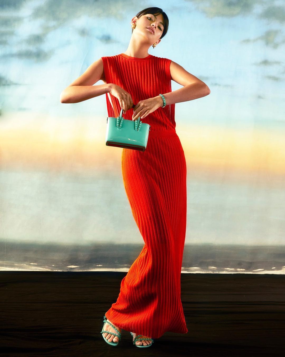 Fashion shot of a woman in an orange dress, holding a turquoise handbag against a painterly background with a blurred ocean effect.