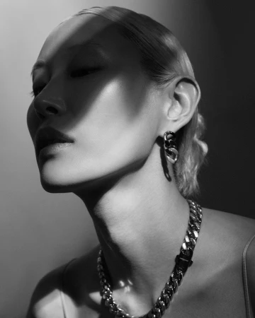 Close-up monochrome portrait of an East Asian woman wearing a chunky chain necklace and matching earrings, captured in dramatic high-contrast light.