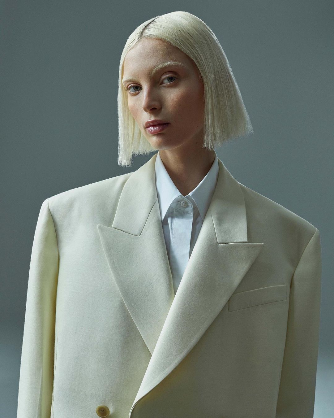 Headshot of a young adult female model with platinum blonde bob haircut and neutral expression, wearing a cream-colored blazer and collared shirt.