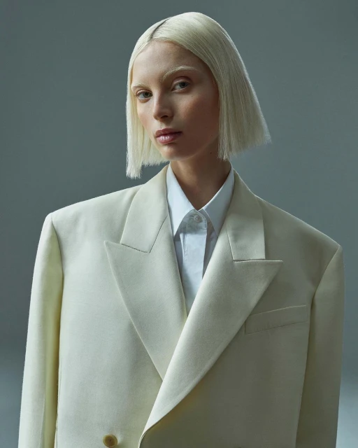 Headshot of a young adult female model with platinum blonde bob haircut and neutral expression, wearing a cream-colored blazer and collared shirt.