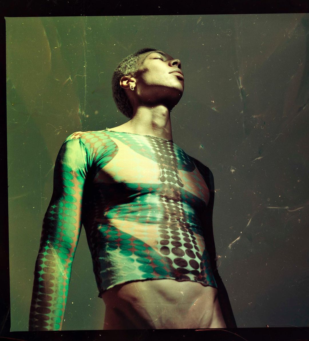 Portrait of a slender adult male with short dyed hair, wearing a patterned mesh shirt, posing in a photo studio with neutral lighting.