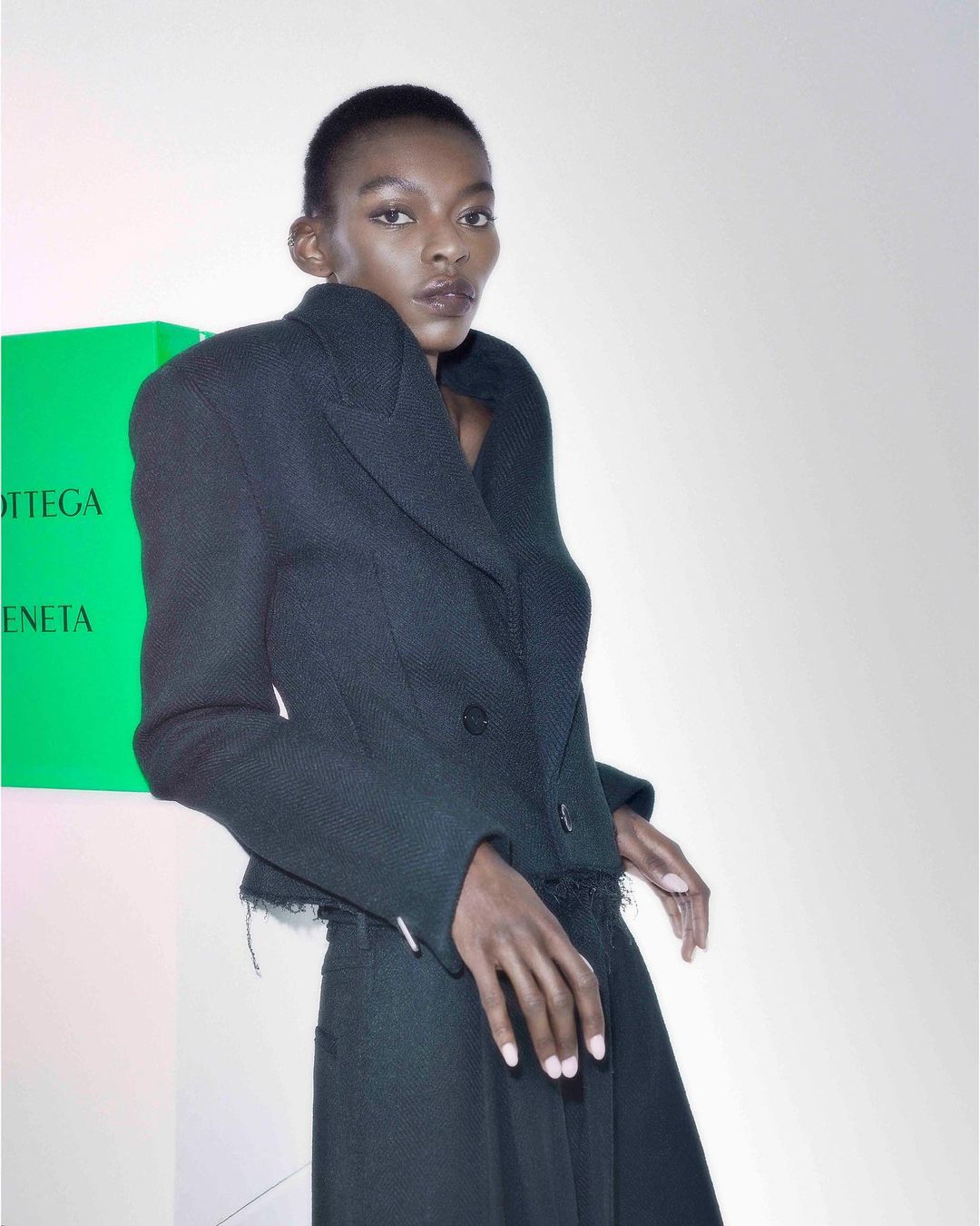 Fashionable woman in a cropped black blazer and long skirt poses indoors against a white backdrop with a green logo visible.