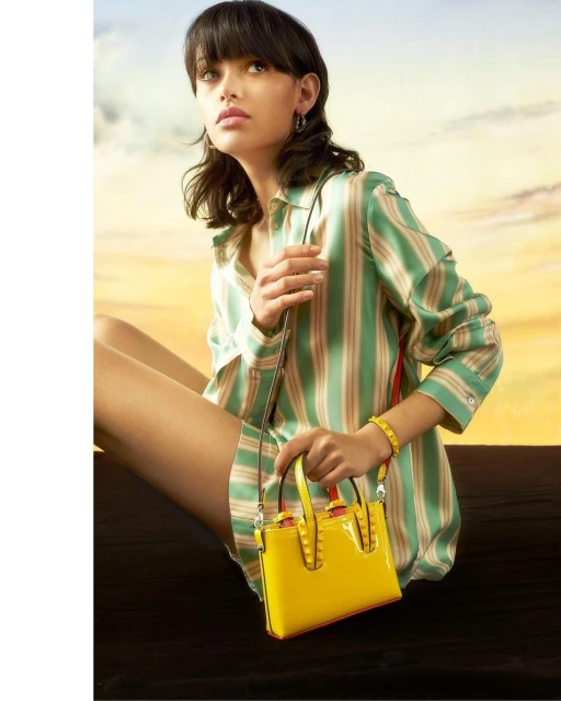 Woman poses with a yellow handbag in a fashion shot. She wears a striped button-down shirt, with a sunset backdrop creating warm lighting.